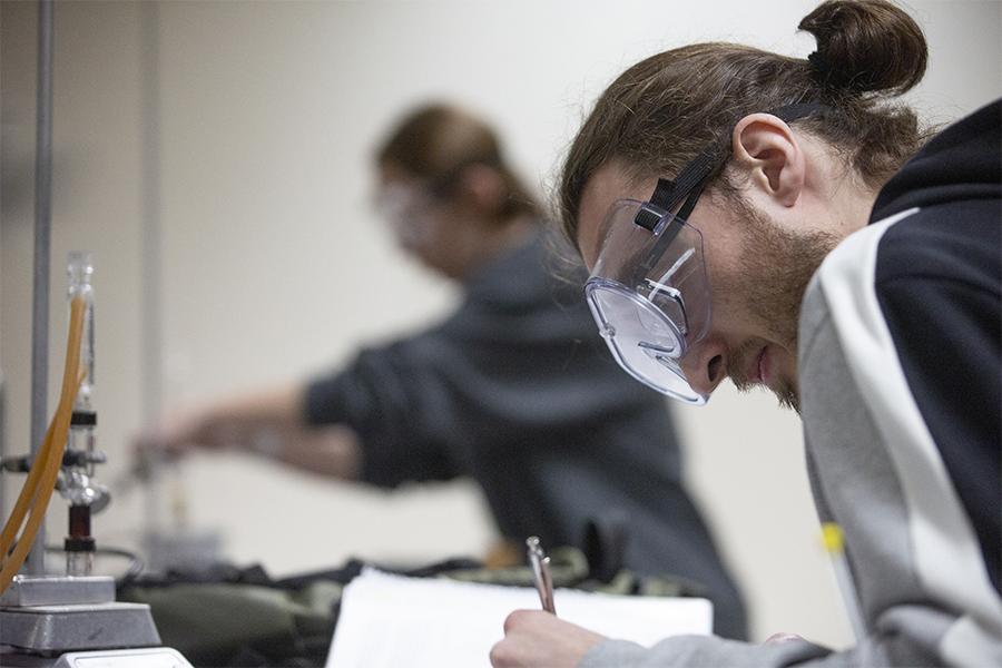 A student wearing safety goggles writes in a notebook.